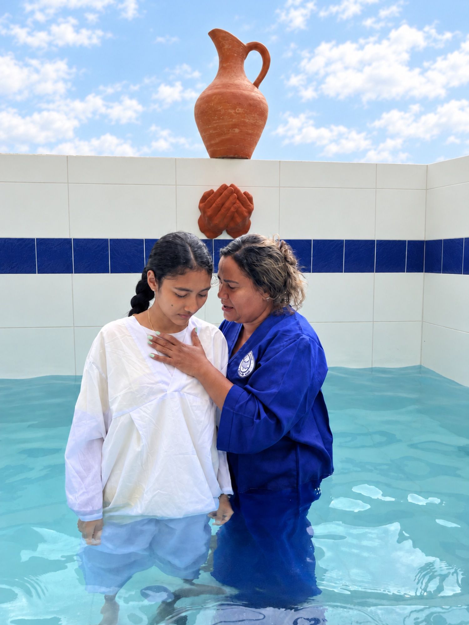 Bautizo en agua: una mujer con túnica azul ora por una joven vestida de blanco en el estanque bautismal